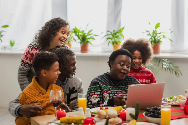 Afrikanische Amerikanerin mittleren Alters spricht während eines Videogesprächs in der Nähe einer fröhlichen Familie und serviert Abendessen — Stockfoto