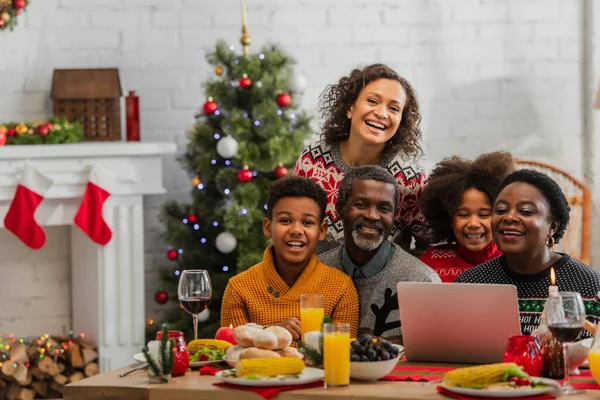 Família afro-americana sorrindo perto de laptop na mesa com jantar de Natal — Fotografia de Stock