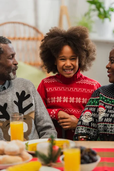 Menina americana africana alegre olhando para a câmera perto de avós sorridentes no jantar de Natal — Fotografia de Stock