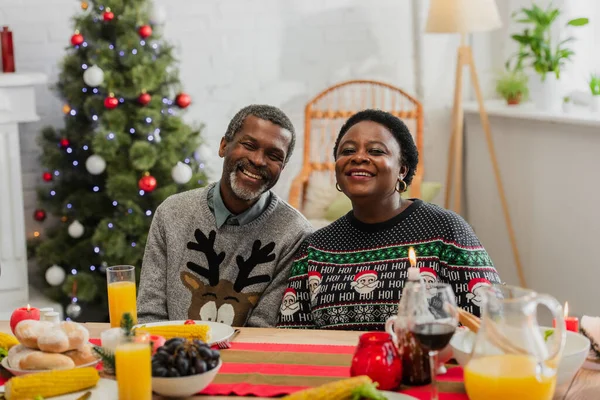 Allegra coppia afro-americana vicino al tavolo servita con cena festiva e albero di Natale su sfondo sfocato — Foto stock