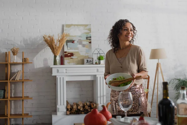 Улыбающаяся африканская американка держит миску со свежим овощным салатом рядом со столом с праздничным ужином — стоковое фото