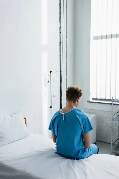 Visão traseira do homem encaracolado em vestido de paciente sentado na cama do hospital — Fotografia de Stock