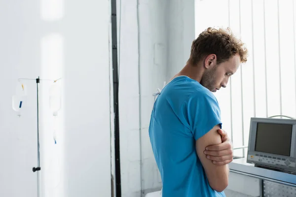 Sad man in patient gown standing with crossed arms in hospital — Stock Photo