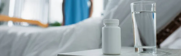 Bouteille avec médicament et verre d'eau sur la table de chevet près du patient flou à l'hôpital, bannière — Photo de stock