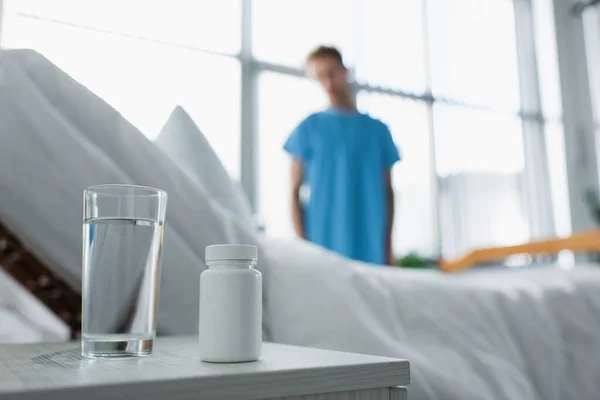 Bottle with medication and glass of water on bedside table near blurred patient in hospital — Stock Photo