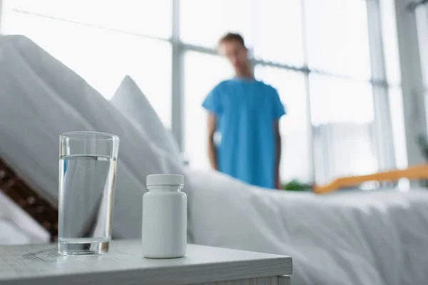 Bottiglia con farmaco e bicchiere d'acqua sul comodino vicino al paziente offuscato in ospedale — Stock Photo