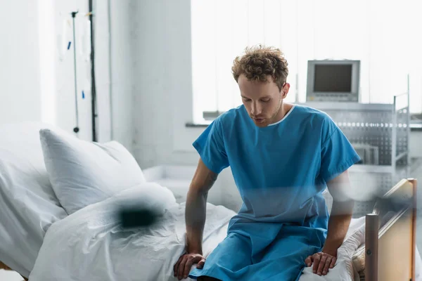 Hombre rizado en bata de paciente sentado en la cama del hospital - foto de stock