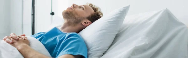 Encaracolado jovem paciente com as mãos apertadas deitado na cama do hospital, banner — Fotografia de Stock