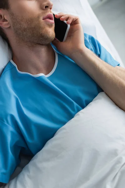 Cropped view of man in patient gown talking on smartphone in hospital bed — Stock Photo