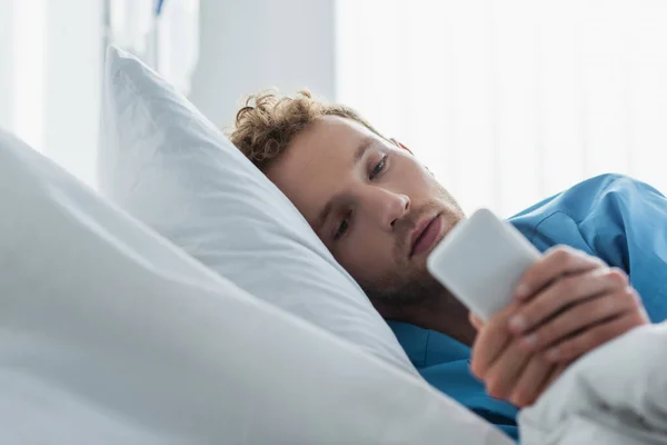 Mensajería paciente rizado en el teléfono inteligente mientras descansa en la cama de hospital - foto de stock