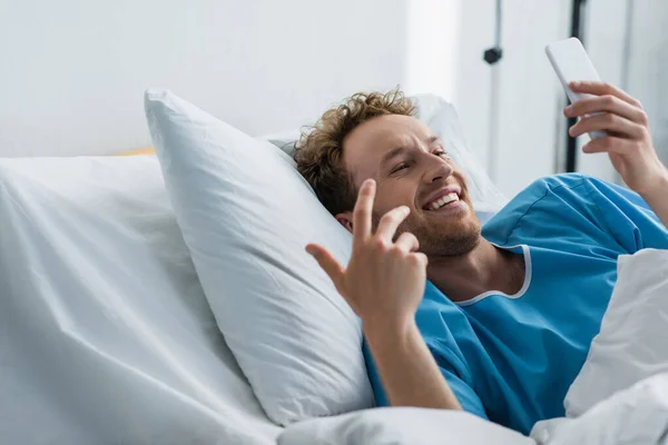 Homme gai en robe de patient ayant appel vidéo sur smartphone à l'hôpital — Photo de stock