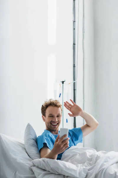 Paciente feliz acenando a mão enquanto tem chamada de vídeo no smartphone no hospital — Fotografia de Stock