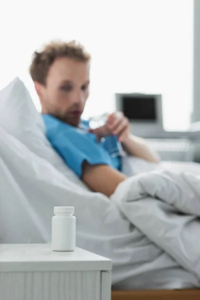 Bottle with medication on bedside table near blurred patient in hospital bed — Stock Photo