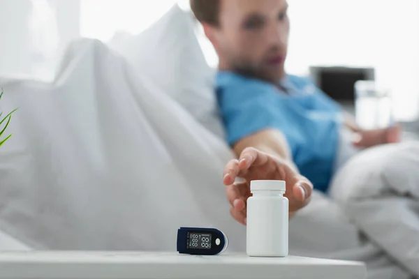 Patient flou atteignant la bouteille avec des médicaments sur la table de chevet près d'un appareil oxymètre — Photo de stock