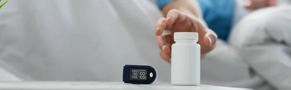 Cropped view of blurred patient reaching bottle with medication on bedside table near oximeter device, banner — Stock Photo