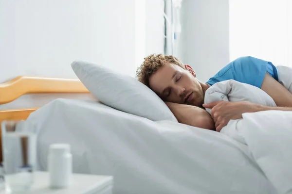 Paziente riccio che dorme in letto d'ospedale vicino a bottiglia sfocata con farmaco e bicchiere d'acqua — Foto stock