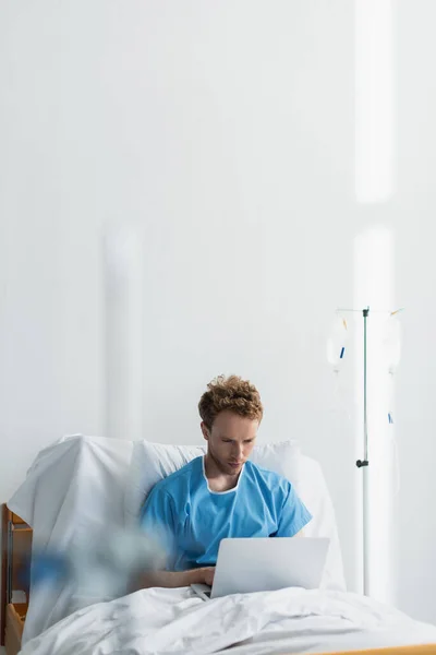 Freelancer em vestido de paciente usando laptop na cama do hospital perto do balcão — Fotografia de Stock