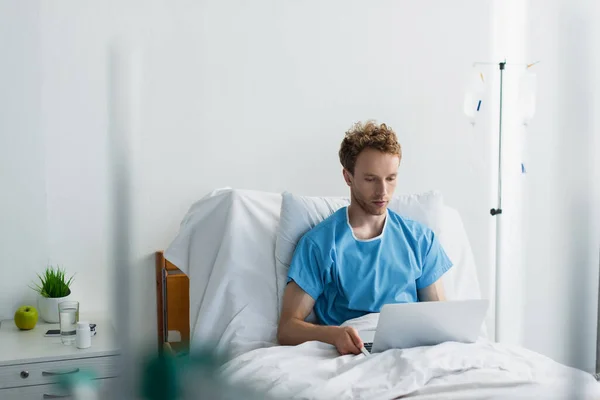 Freelancer em vestido de paciente usando laptop na cama do hospital — Fotografia de Stock