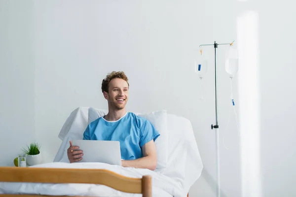 Freelancer alegre em vestido de paciente usando laptop na cama do hospital — Fotografia de Stock