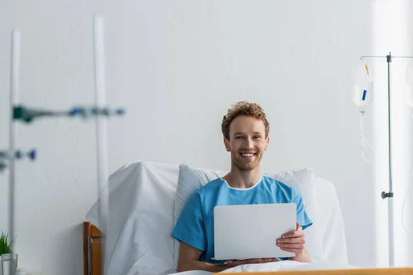 Freelancer alegre en bata de paciente usando el ordenador portátil en la cama del hospital - foto de stock