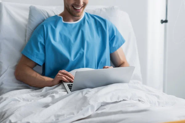 Vista recortada de freelancer feliz en el vestido del paciente usando el ordenador portátil en la cama del hospital - foto de stock