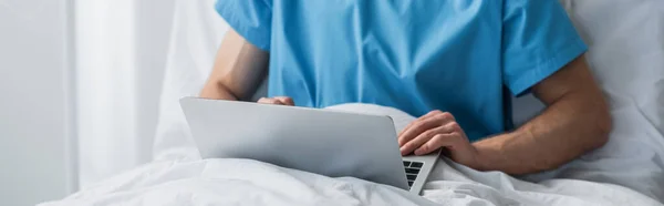 Vista cortada de freelancer em vestido de paciente usando laptop na cama do hospital, banner — Fotografia de Stock