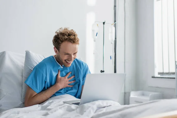 Homem feliz em vestido de paciente rindo ao usar laptop na cama do hospital — Fotografia de Stock