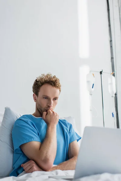 Curly man in patient gown looking at laptop in hospital bed — Stock Photo