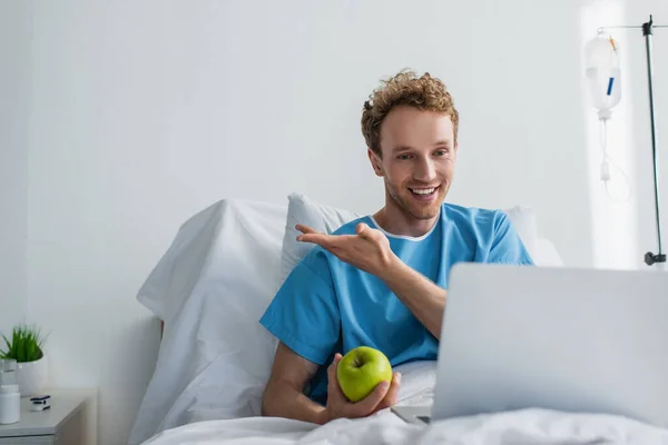 Paciente alegre señalando con la mano mientras tiene videollamada y la celebración de manzana en el hospital - foto de stock