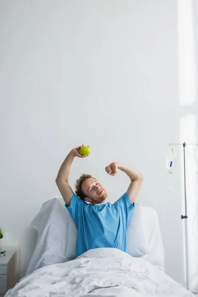 Задоволений чоловік з закритими очима розтягується в лікарняному ліжку і тримає яблуко — стокове фото