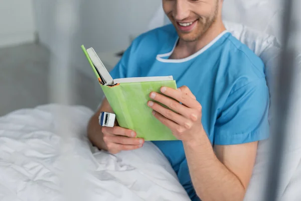 Cropped view of cheerful patient with oximeter on finger reading book in hospital bed — Stock Photo