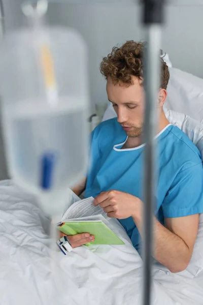 Patient avec oxymètre sur le livre de lecture des doigts dans un lit d'hôpital près d'un compteur de gouttes flou — Photo de stock