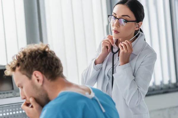 Brünette Ärztin mit Brille untersucht verschwommene und kranke Patientin mit Stethoskop — Stockfoto