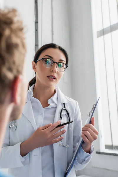 Doctor in glasses holding pen and clipboard while looking at blurred patient — Stock Photo