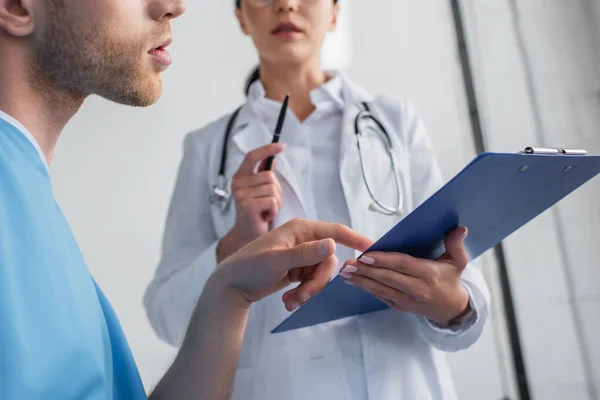 Cropped view of patient pointing at clipboard in hands of blurred doctor in clinic — Stock Photo