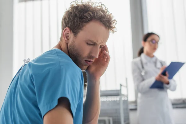 Curly patient suffering from headache near blurred doctor in hospital — Stock Photo