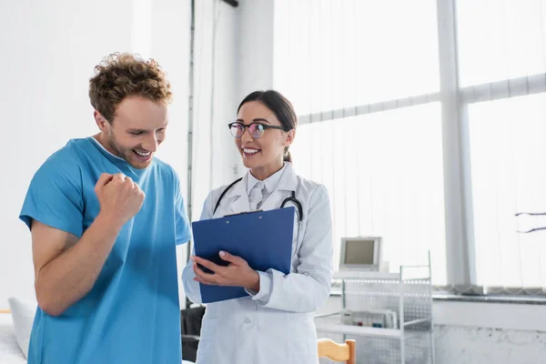 Heureux et brunette médecin avec presse-papiers près patient joyeux — Photo de stock