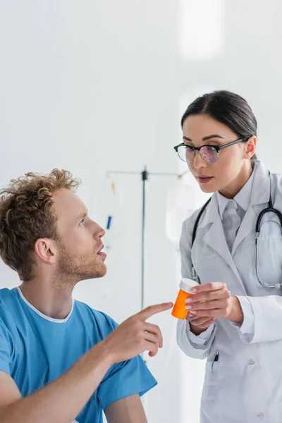 Lockiger Patient zeigt in weißem Kittel und Brille in der Nähe des Arztes auf Flasche mit Medikamenten — Stockfoto