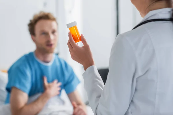 Врач в белом халате держит бутылку с лекарствами рядом с размытым пациентом — стоковое фото