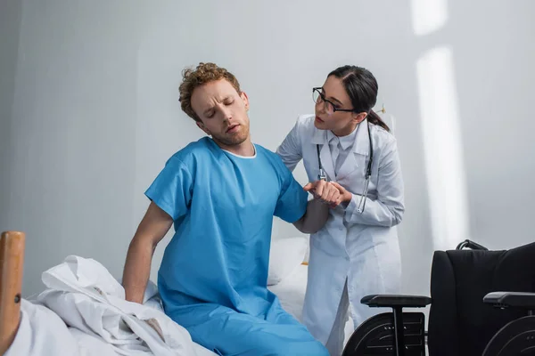 Médico en gafas ayudando al paciente a levantarse de la cama del hospital cerca de la silla de ruedas - foto de stock