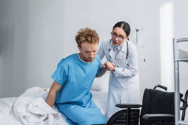 Médico em óculos ajudando paciente encaracolado se levantar da cama do hospital perto de cadeira de rodas — Fotografia de Stock