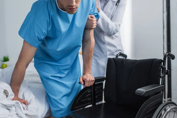 Visão recortada do médico de casaco branco ajudando o paciente a levantar-se da cama do hospital perto da cadeira de rodas — Fotografia de Stock