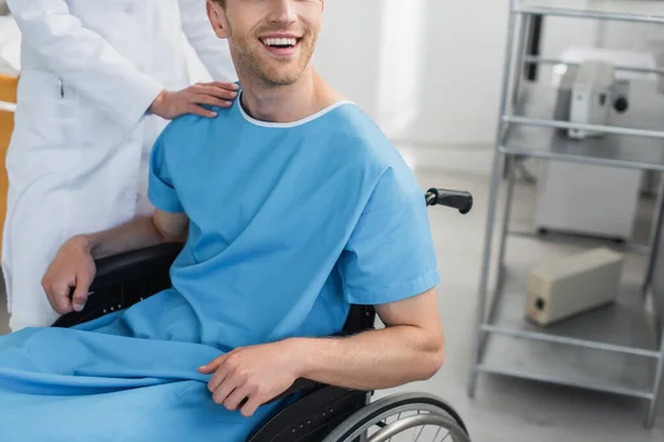 Обрезанный вид врача в белом халате, стоящего рядом с веселым инвалидом в инвалидном кресле — стоковое фото