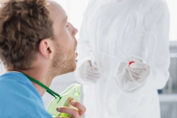 Doctor in hazmat suit and latex gloves holding swab near patient with open mouth — Stock Photo