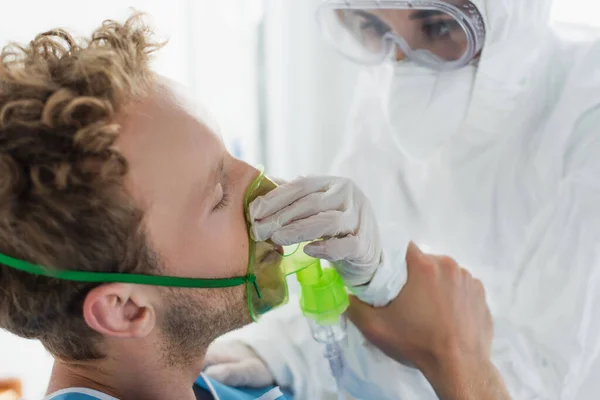 Médico en traje de materiales peligrosos ajustando la máscara de oxígeno en el paciente en el hospital - foto de stock