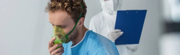 Kranker Patient atmet Sauerstoffmaske in der Nähe verschwommener Arzt in persönlicher Schutzausrüstung, Banner — Stockfoto