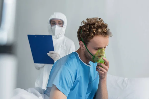 Doente respirando em máscara de oxigênio perto médico borrado em equipamento de proteção individual segurando prancheta — Fotografia de Stock