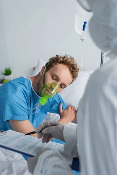 Doctor borroso en traje de materiales peligrosos sujetando el portapapeles y la pluma cerca del paciente con máscara de oxígeno - foto de stock