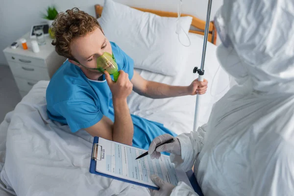Visão de alto ângulo do médico em terno hazmat segurando prancheta e caneta perto do paciente em máscara de oxigênio — Fotografia de Stock