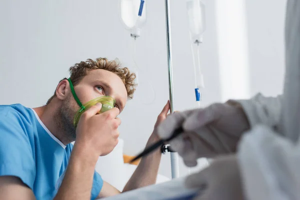 Paziente in maschera di ossigeno guardando medico offuscata con appunti e penna — Foto stock